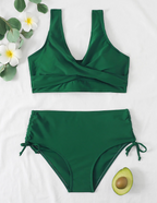 Maillot de bain vert deux pièces avec haut croisé et culotte taille haute, décoré de fleurs blanches et demi-avocat sur fond blanc.