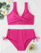 Maillot de bain deux pièces rose vif avec haut croisé et culotte à cordons, posé sur fond blanc avec fleurs et demi-avocat.