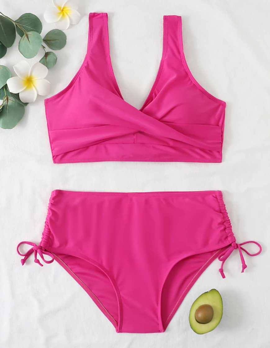 Maillot de bain deux pièces rose vif avec haut croisé et culotte à cordons, posé sur fond blanc avec fleurs et demi-avocat.