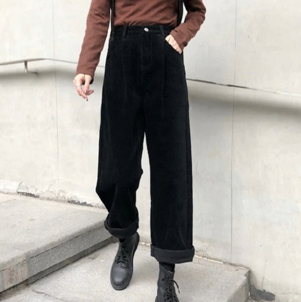Pantalon ample noir en velours côtelé, porté avec un pull marron et des bottes noires, style urbain moderne sur fond de mur gris.