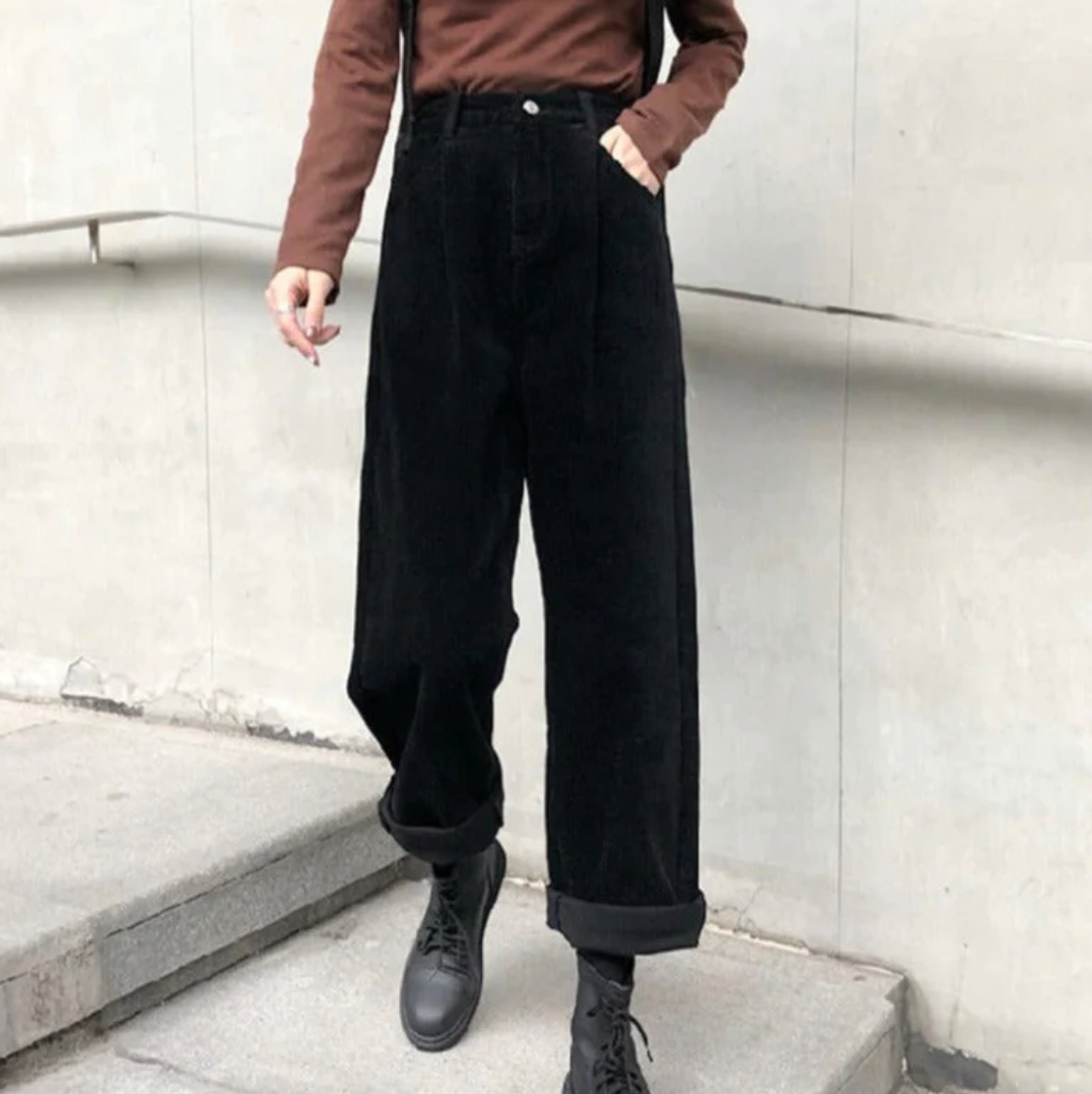 Pantalon ample noir en velours côtelé, porté avec un pull marron et des bottes noires, style urbain moderne sur fond de mur gris.