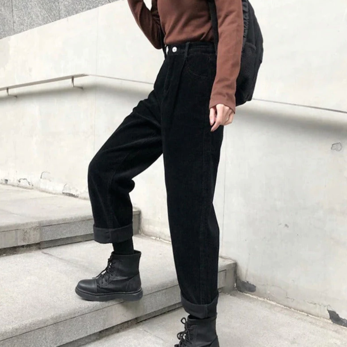 Jeune femme en pantalon noir ample et bottes noires montant des escaliers en béton, portant un pull marron et un sac à dos noir.