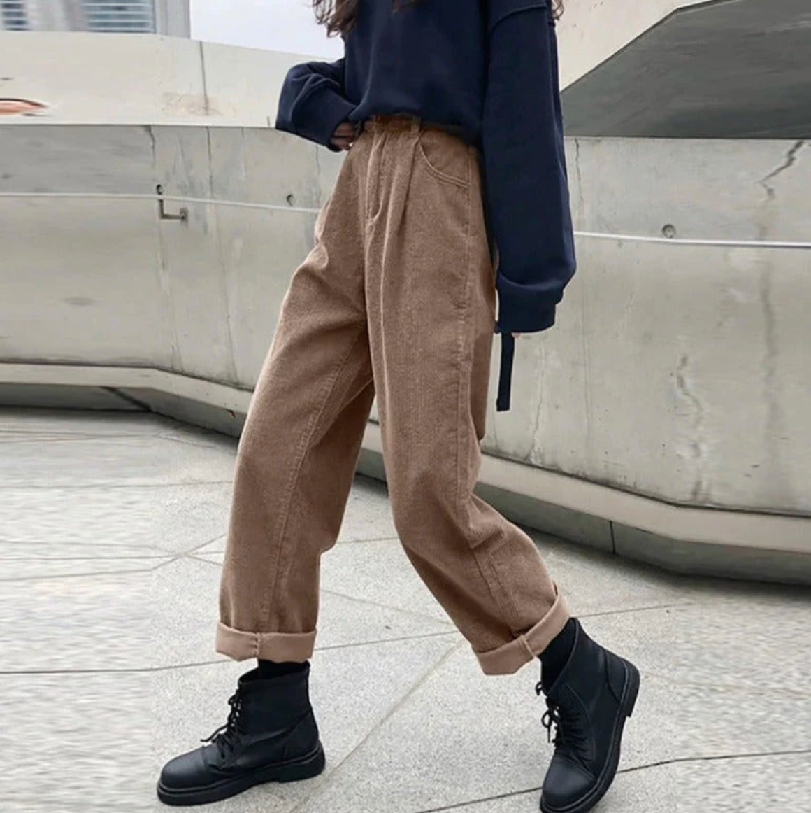 Jeune femme portant un pantalon beige ample et un pull bleu marine, chaussures noires, posant sur un trottoir urbain moderne. Mode décontractée tendance.