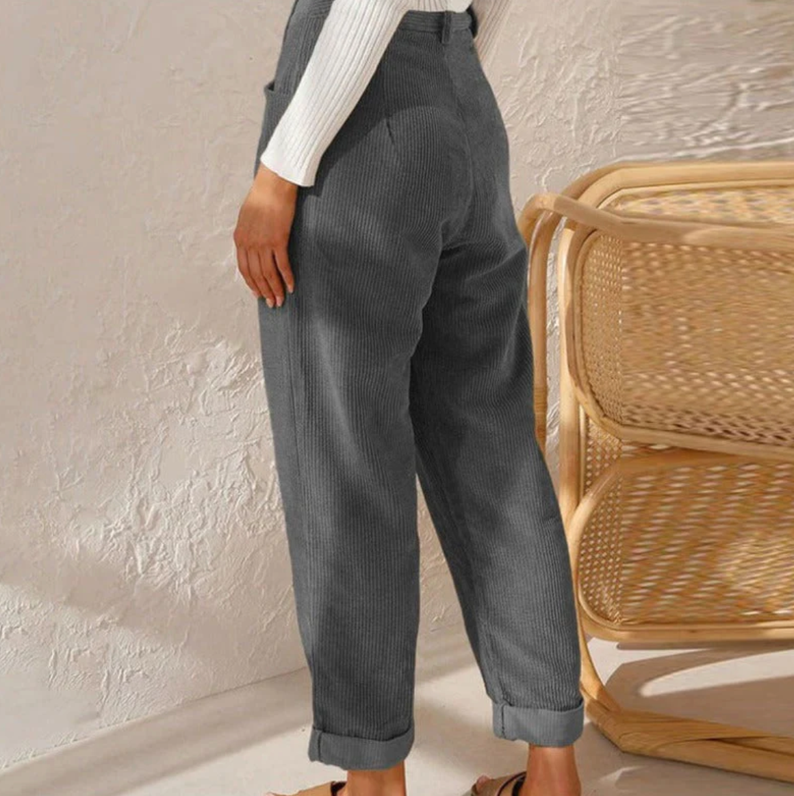 Pantalon en velours côtelé gris pour femme, style décontracté, avec ourlets retroussés, associé à un pull blanc, près d'une chaise en rotin.