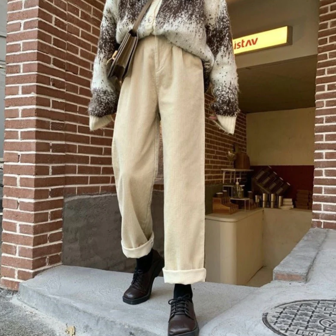 Personne portant un pull ample à motifs et un pantalon beige, debout devant un mur de briques. Mode automne, style décontracté, chaussures noires.