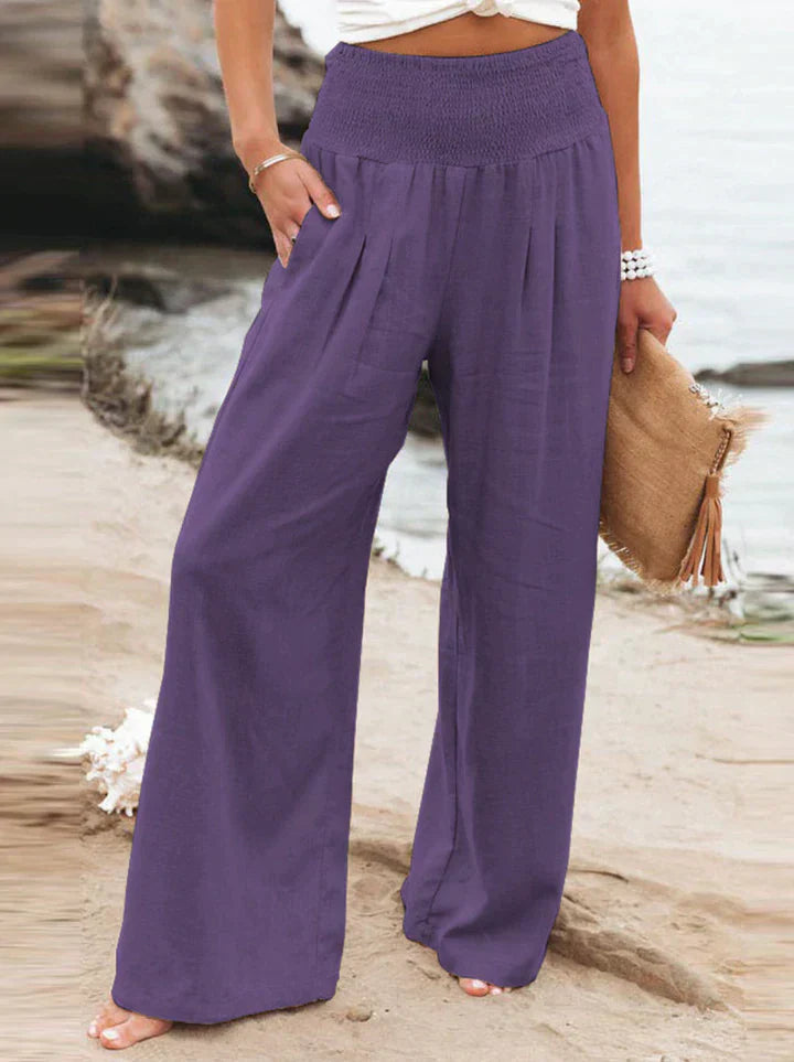 Pantalon large violet femme, taille haute, plage en arrière-plan, style décontracté, mode estivale, accessoire sac en paille, tenue élégante.