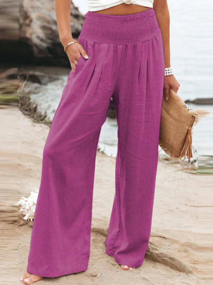 Pantalon large femme violet, taille haute, en lin, porté sur une plage. Mode estivale, tenue décontractée, accessoire sac en paille.