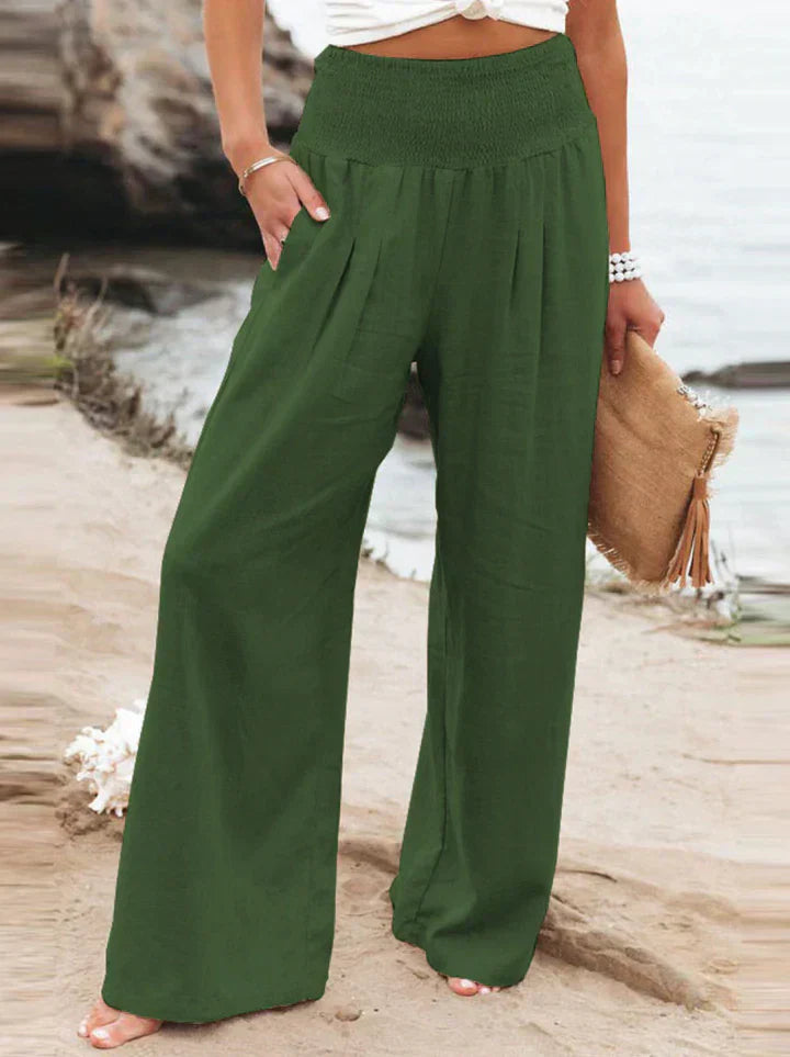 Pantalon large vert pour femme, taille haute, style décontracté, plage en arrière-plan, accessoire sac en paille, mode estivale tendance.