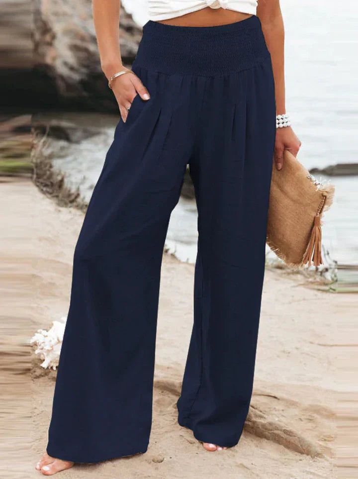 Pantalon large bleu marine pour femme, style décontracté, plage en arrière-plan, accessoire sac en paille, mode estivale élégante.