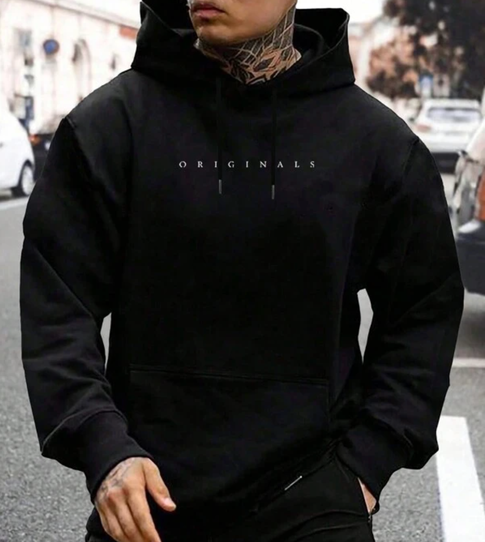 Homme portant un sweat à capuche noir "Originals" avec tatouages visibles, marchant en ville. Mode urbaine, style streetwear, tendance masculine.