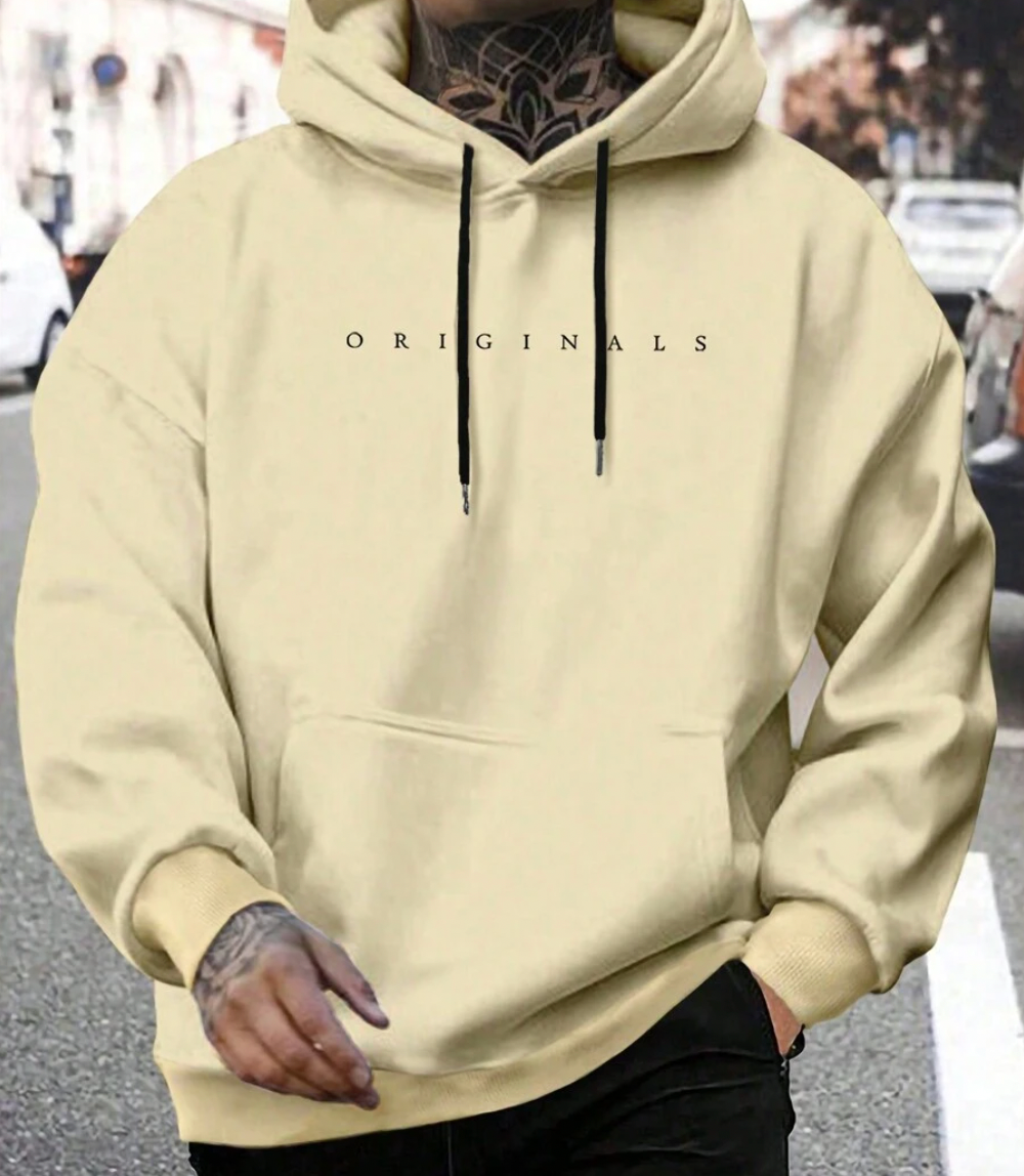 Homme portant un sweat à capuche beige "ORIGINALS" dans la rue, tatouages visibles, mode urbaine, style décontracté, tendance automne-hiver.