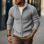 Homme portant un pull zippé gris en maille côtelée, manches longues, sur un t-shirt blanc, avec pantalon marron, style décontracté chic.