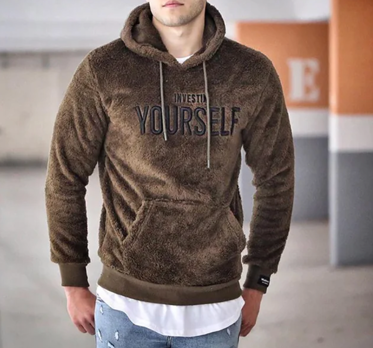 Homme portant un sweat à capuche marron en peluche avec l'inscription "Invest in Yourself", dans un parking. Mode décontractée, tendance automne.