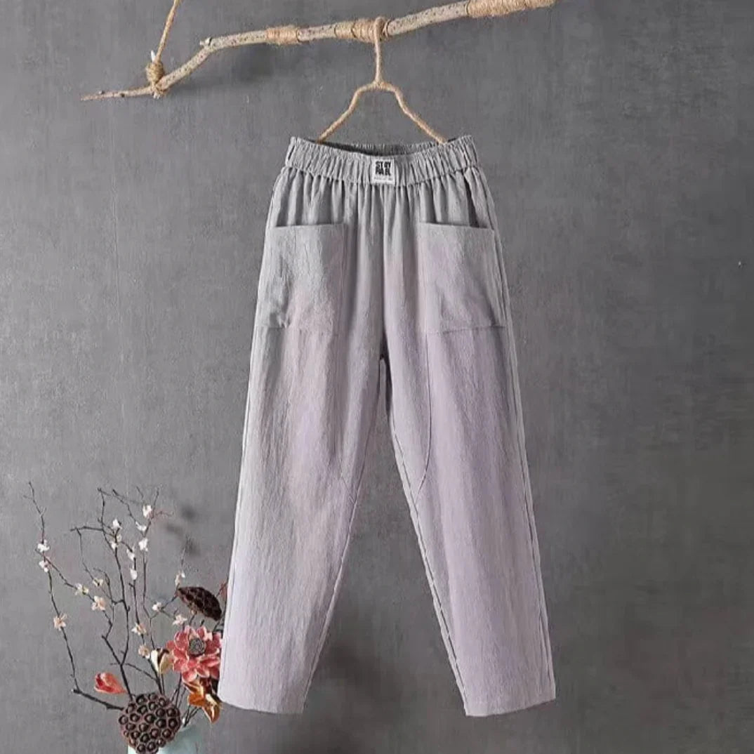 Pantalon en lin gris suspendu à une branche, style décontracté, poches avant, fond gris, décoration florale en bas à gauche.
