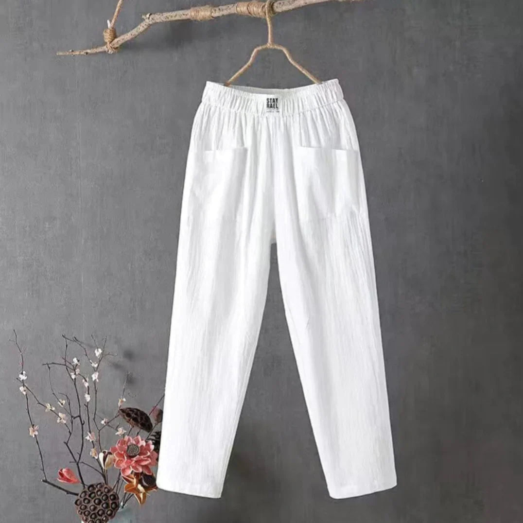 Pantalon blanc en lin suspendu sur un cintre en bois, fond gris, décor floral en bas à gauche. Mode éthique, vêtements légers, style décontracté.