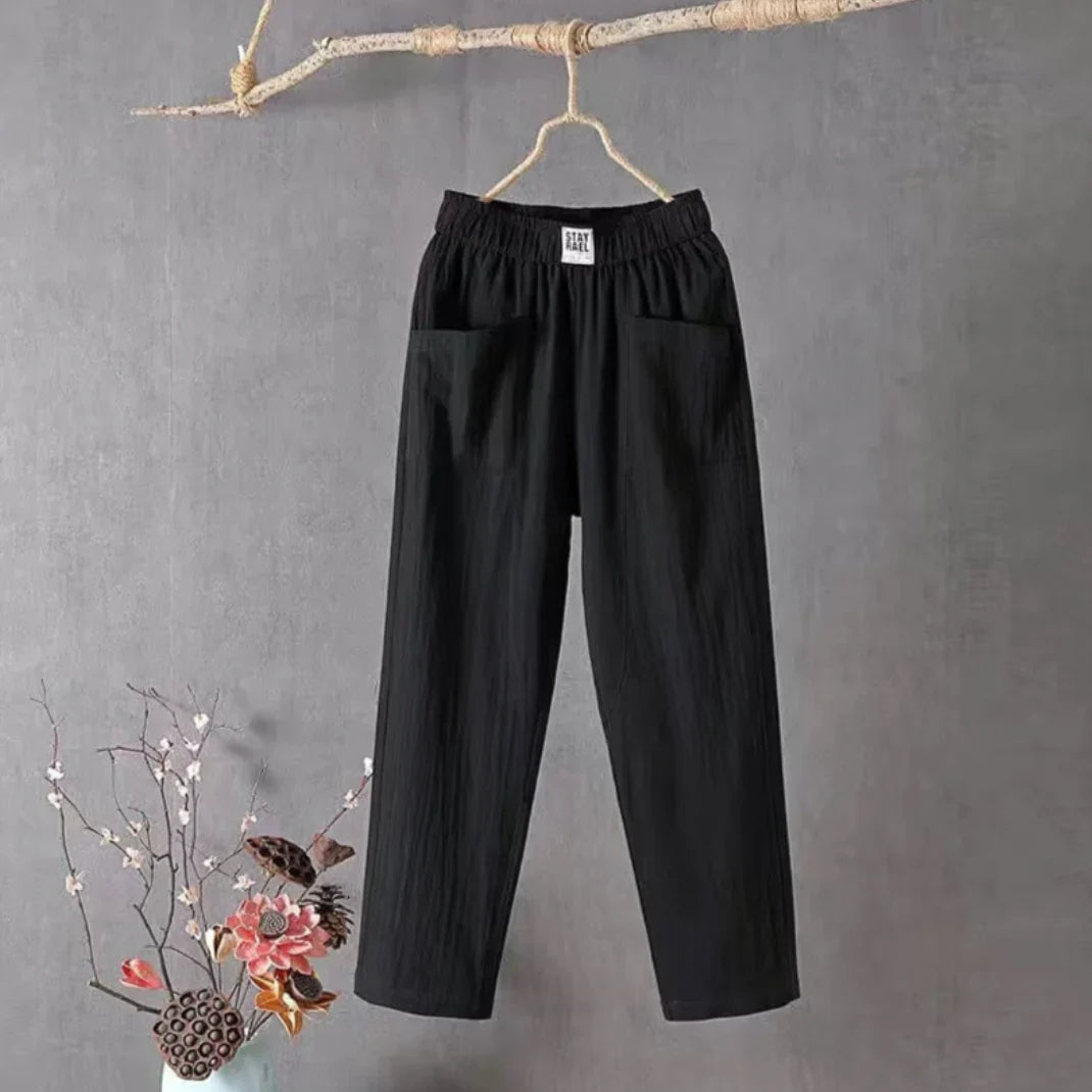 Pantalon noir ample suspendu sur une branche, avec poches avant, style décontracté. Fond gris, décor floral en bas à gauche. Mode minimaliste.