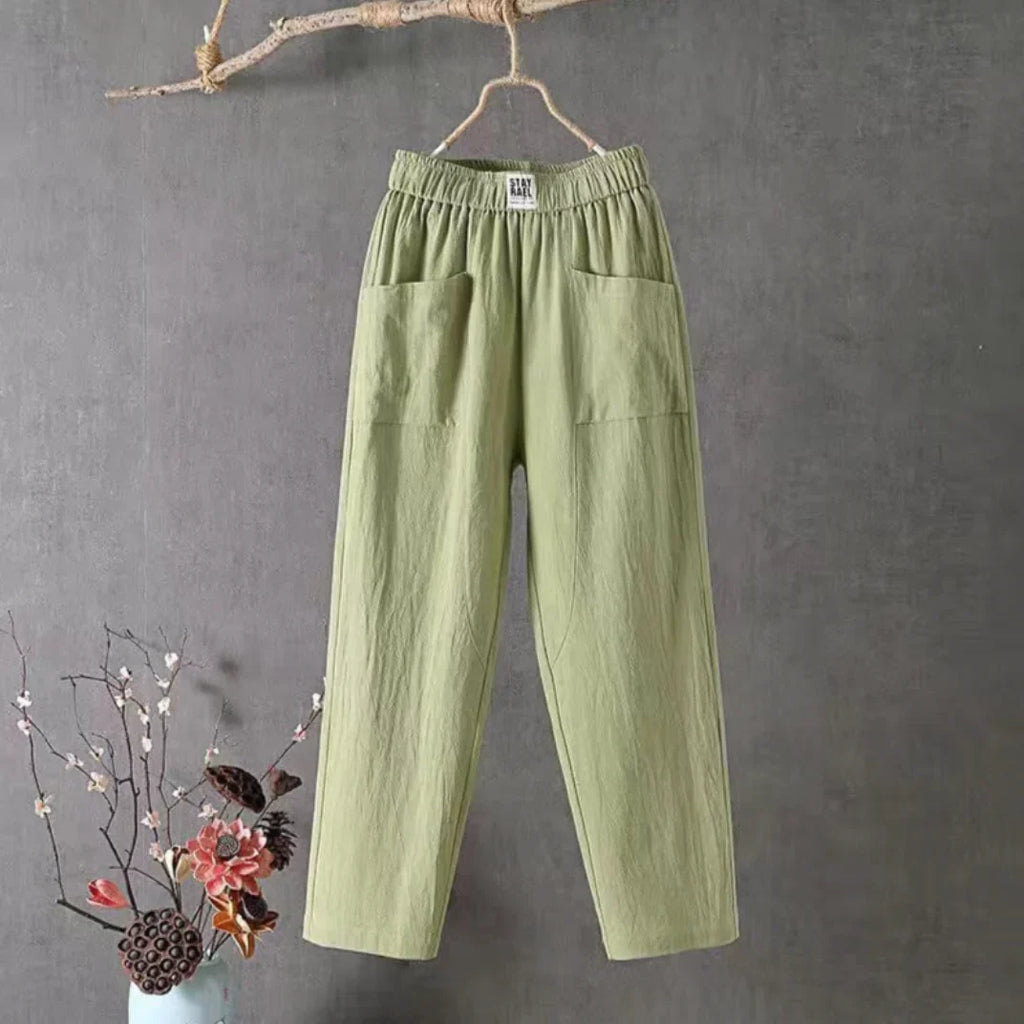 Pantalon vert en lin suspendu, poches avant, fond gris, branche décorative, vase avec fleurs, style décontracté, mode éco-responsable.