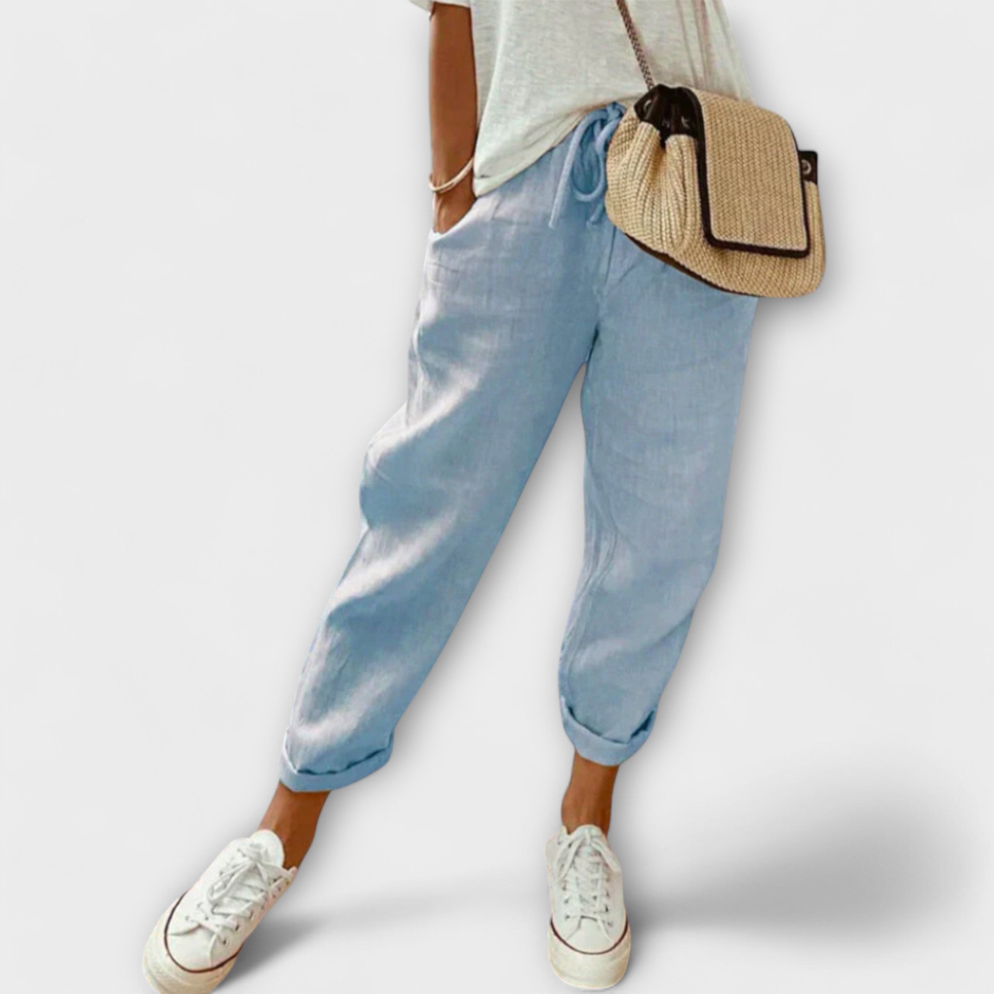 Personne portant un pantalon en lin bleu clair, t-shirt blanc, baskets blanches et sac en osier. Mode décontractée, style estival, tenue confortable.