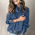Femme souriante portant un pull bleu à col roulé avec boutons décoratifs, cheveux ondulés, boucles d'oreilles dorées, bracelet, jeans décontractés.