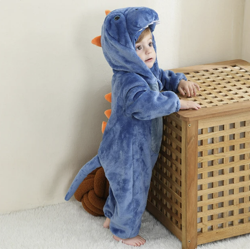 Enfant en grenouillère dinosaure bleue avec capuche, debout près d'un meuble en bois. Costume bébé dinosaure, pyjama chaud et confortable.