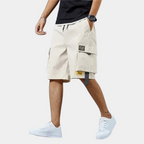 Homme portant un short cargo beige, t-shirt noir et baskets blanches. Mode décontractée, style urbain, tenue estivale pour homme.