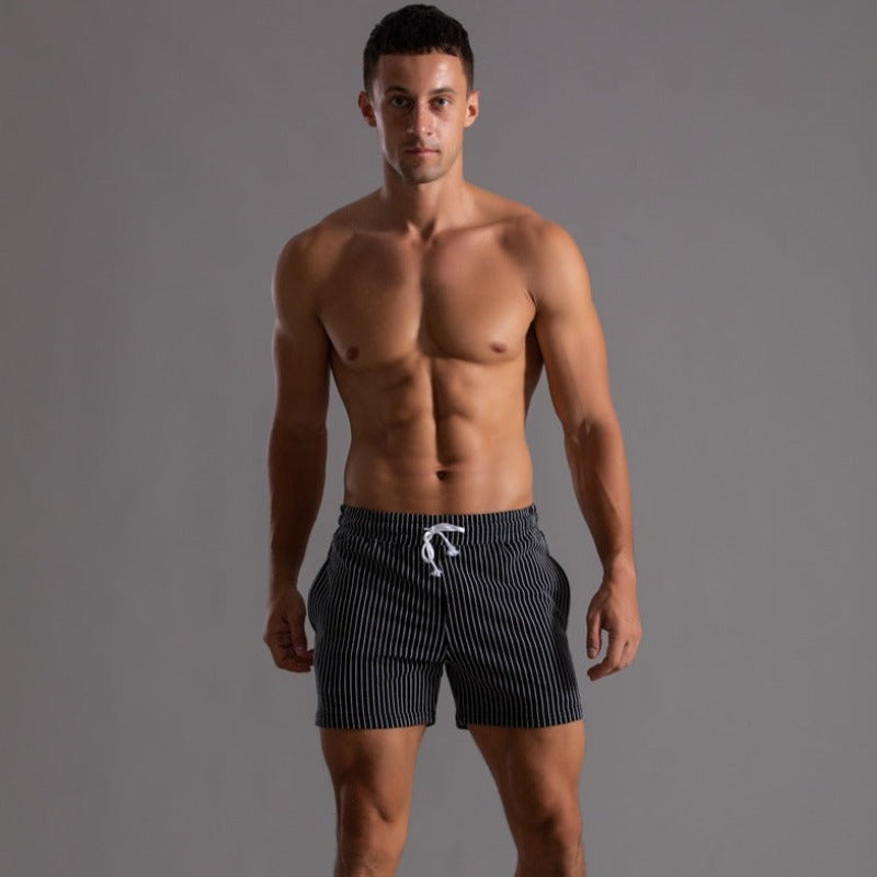 Homme en short de bain rayé noir et blanc, torse nu, sur fond gris. Mode masculine, maillot de bain tendance, été, fitness.