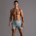 Homme musclé portant un short de bain rayé vert clair, debout sur fond gris. Mode masculine, maillot de bain tendance, été.