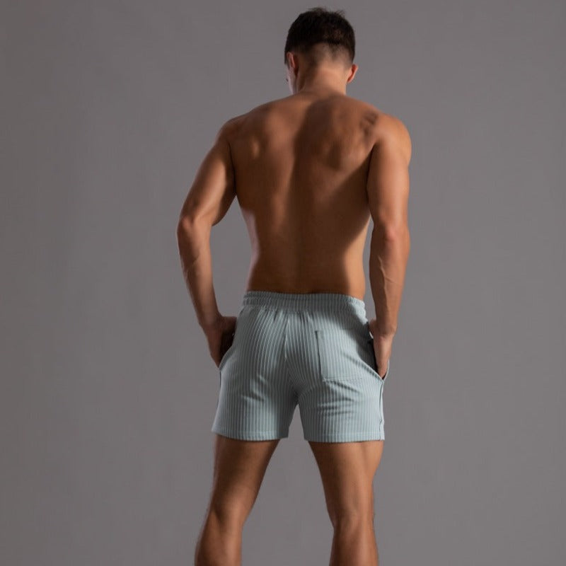 Homme musclé de dos portant un short rayé bleu clair, torse nu, sur fond gris. Mode masculine, vêtements d'été, style décontracté.