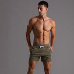 Homme musclé portant un short de bain rayé vert, posant sur fond gris. Mode masculine, maillot de bain tendance, style estival.