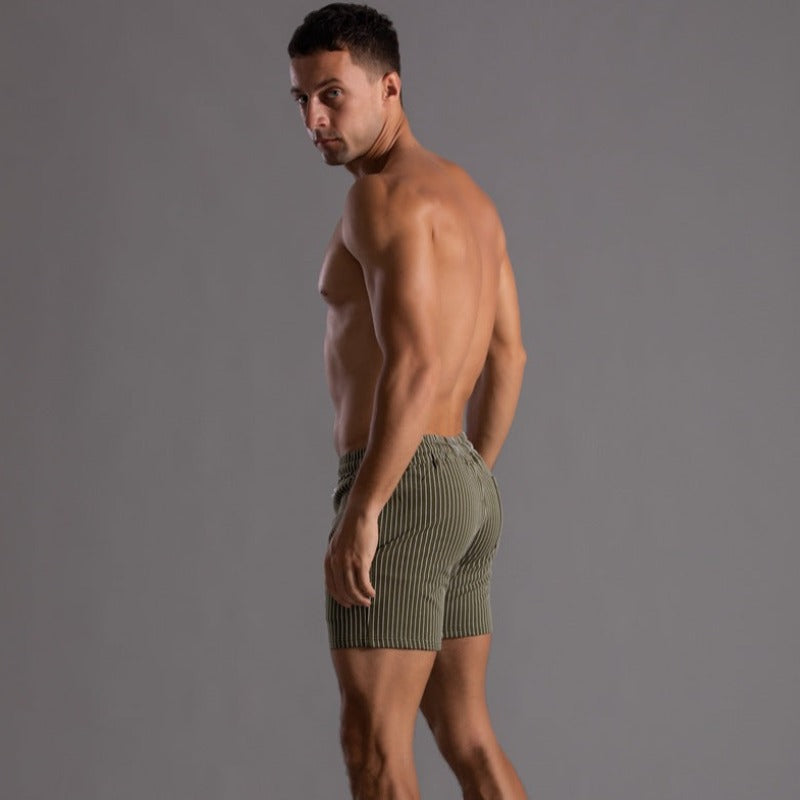 Homme musclé portant un short rayé vert olive, posant de profil sur fond gris. Mode masculine, vêtements d'été, style décontracté.