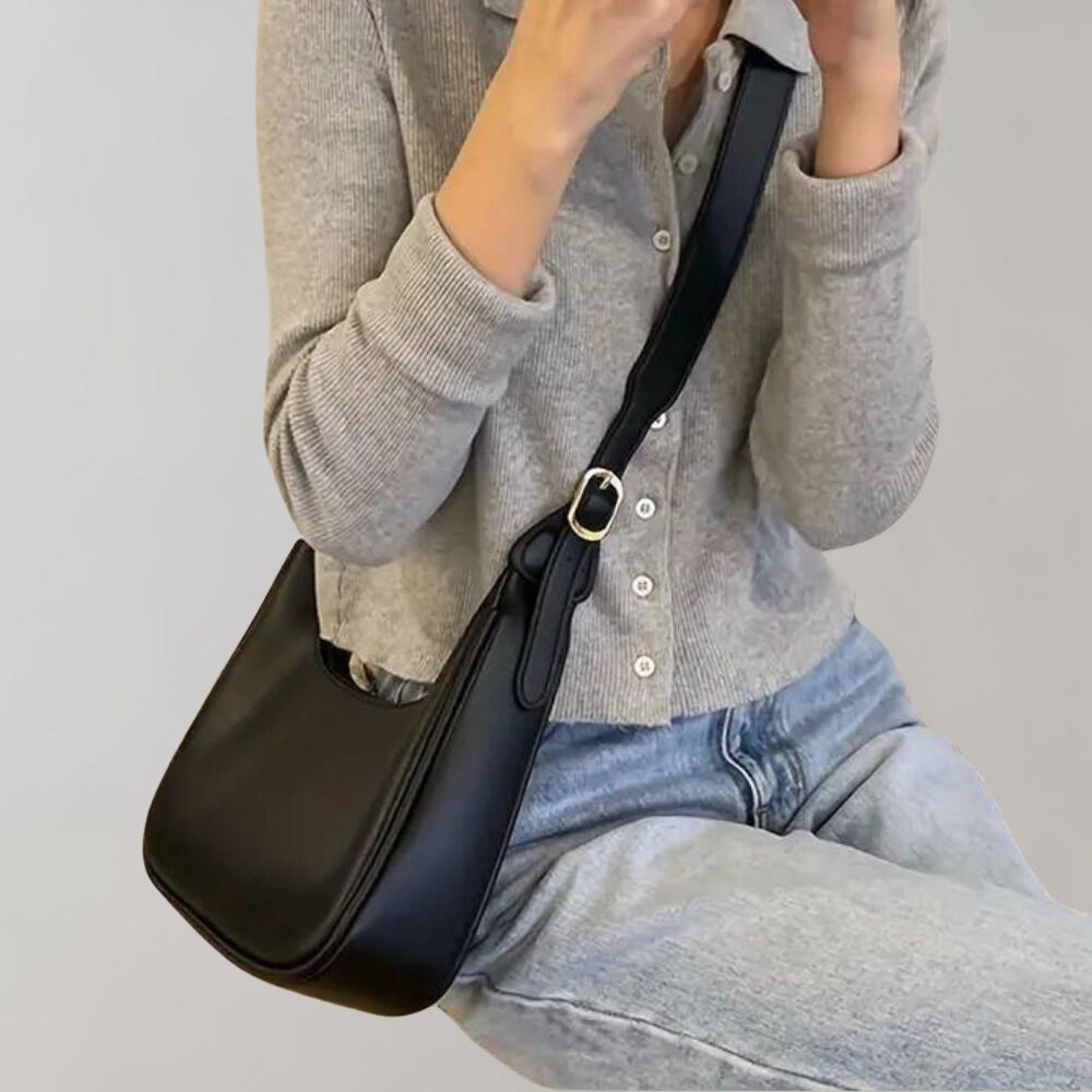 Femme portant un sac à bandoulière noir en cuir, tenue décontractée avec cardigan gris et jean. Mode féminine élégante et moderne.