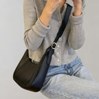 Femme portant un sac à bandoulière noir en cuir, tenue décontractée avec cardigan gris et jean. Mode féminine élégante et moderne.