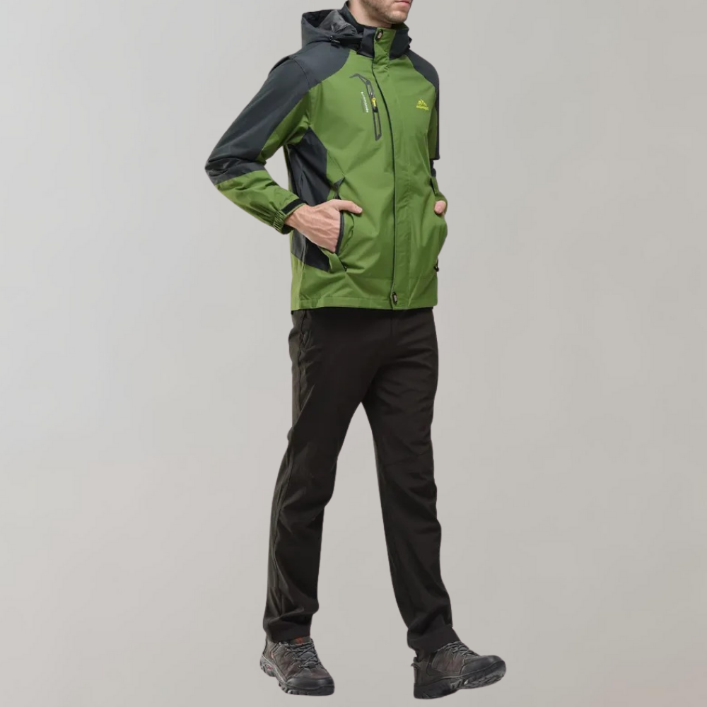 Homme portant une veste imperméable verte et noire, pantalon noir, chaussures de randonnée. Tenue outdoor pour activités en plein air, randonnée.