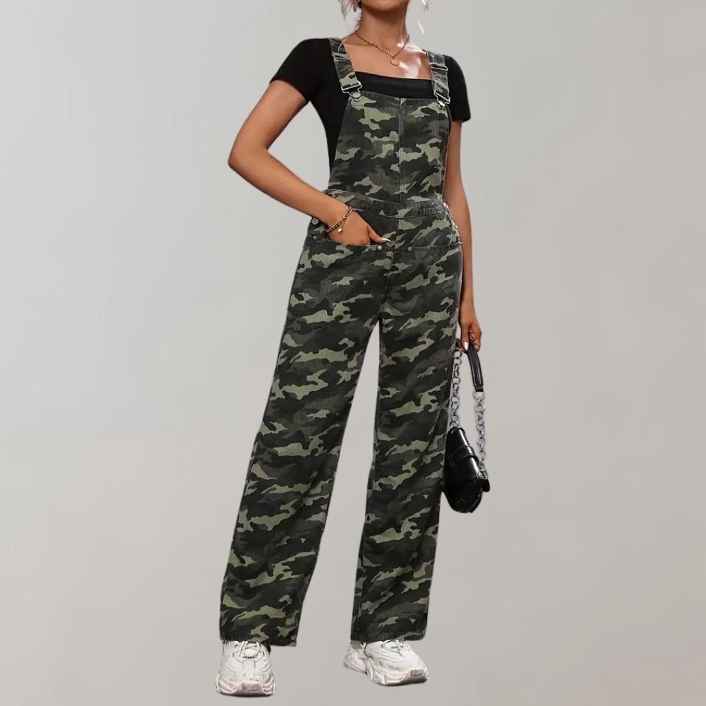 Femme portant une salopette camouflage verte avec un t-shirt noir et des baskets blanches, tenant un sac à main noir. Mode urbaine tendance.