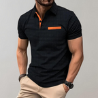 Homme portant un polo noir à motifs avec détails orange, manches courtes, associé à un pantalon beige. Mode masculine élégante et moderne.