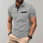 Homme portant un polo gris texturé avec poche noire, manches courtes, associé à un pantalon beige. Mode masculine élégante et décontractée.