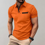 Homme portant un polo orange à manches courtes avec poche noire, style décontracté, sur fond gris. Mode masculine, tenue estivale élégante.