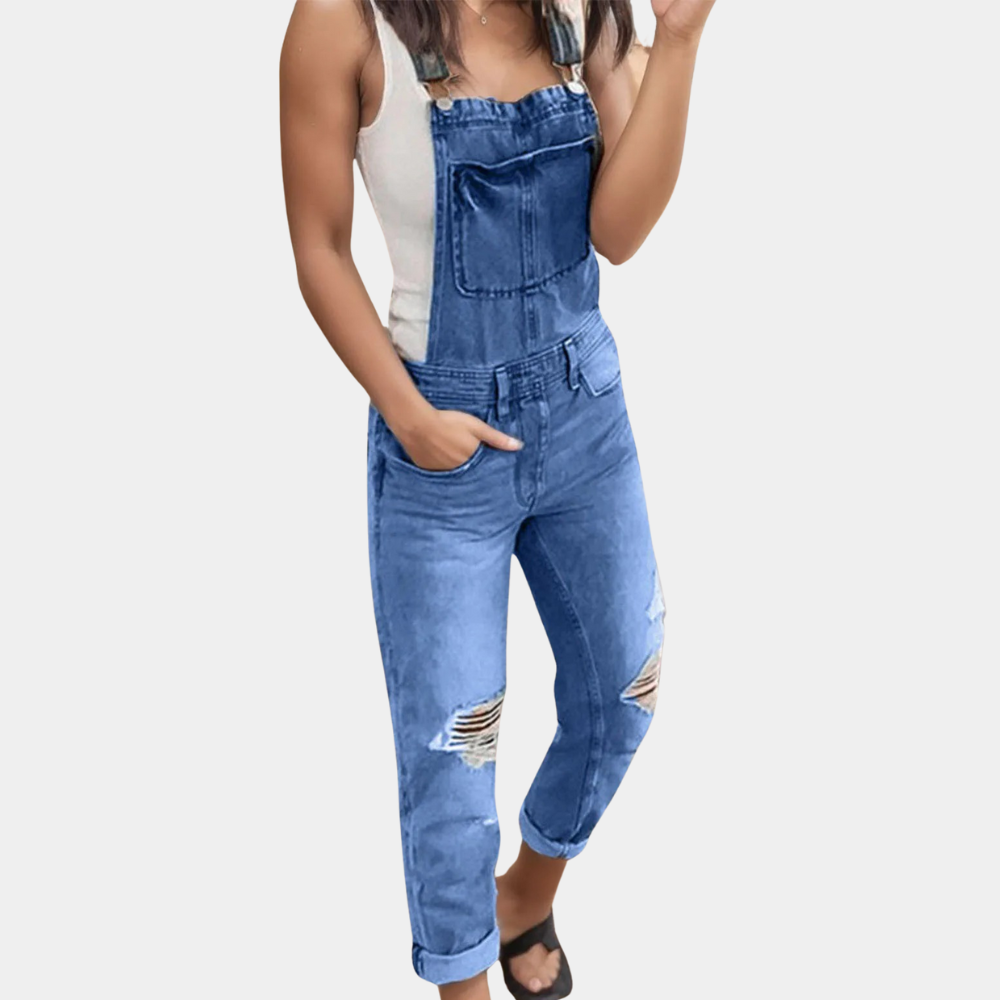 Femme portant une salopette en jean bleu déchiré, associée à un débardeur blanc et des sandales noires. Mode décontractée tendance.