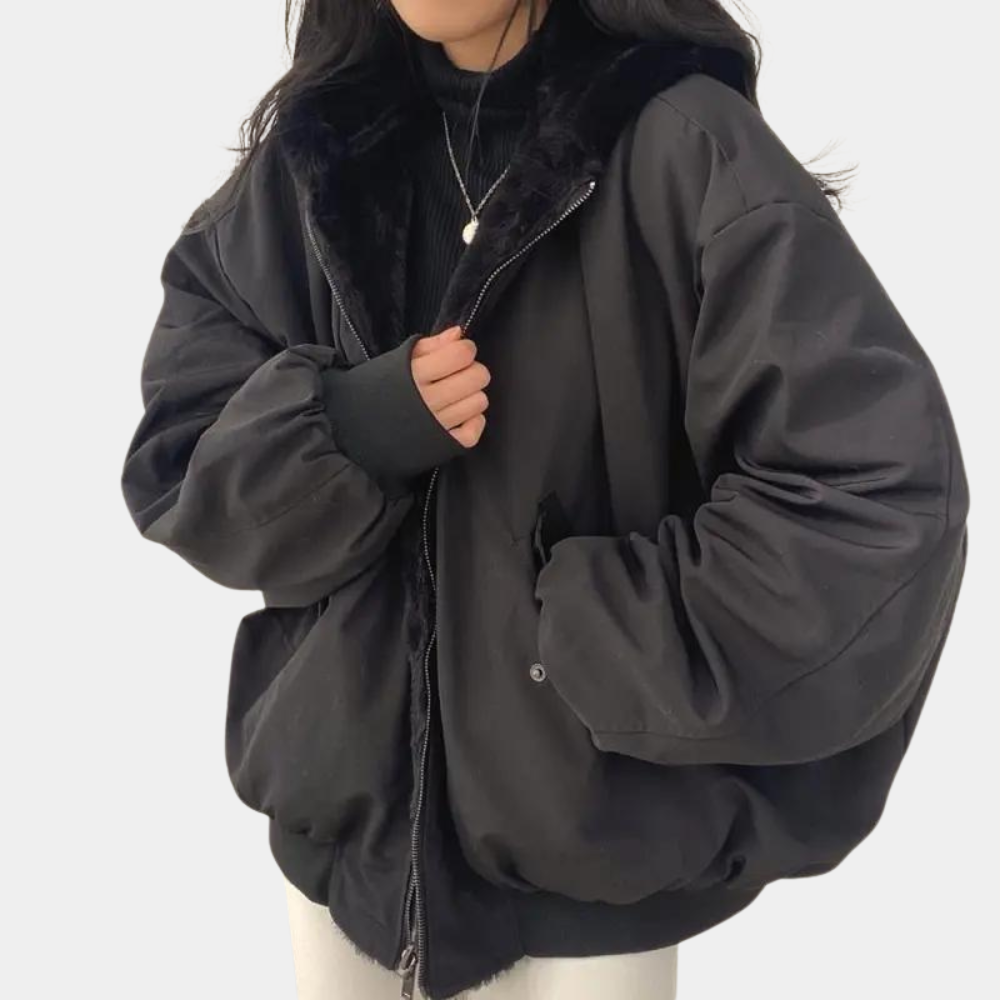 Femme portant une veste noire oversize avec col en fausse fourrure, fermée par une fermeture éclair, sur un pull noir. Mode hiver tendance.