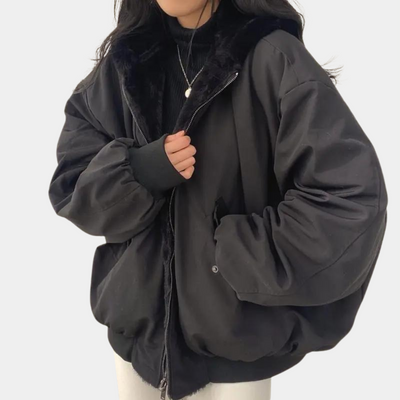 Femme portant une veste noire oversize avec col en fausse fourrure, fermée par une fermeture éclair, sur un pull noir. Mode hiver tendance.