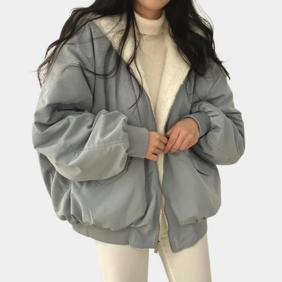 Femme portant une veste oversize grise avec doublure en fourrure blanche, pull beige et pantalon blanc. Mode hiver, style décontracté, tendance féminine.