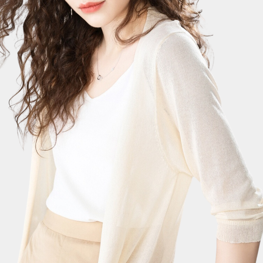 Femme aux cheveux bouclés portant un cardigan beige léger, t-shirt blanc et pantalon beige. Mode féminine élégante et décontractée.