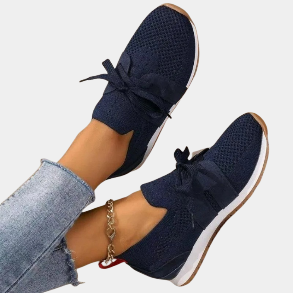 Chaussures de sport bleu marine pour femme, design en maille respirante, semelle blanche, lacets, portées avec un jean et un bracelet de cheville argenté.