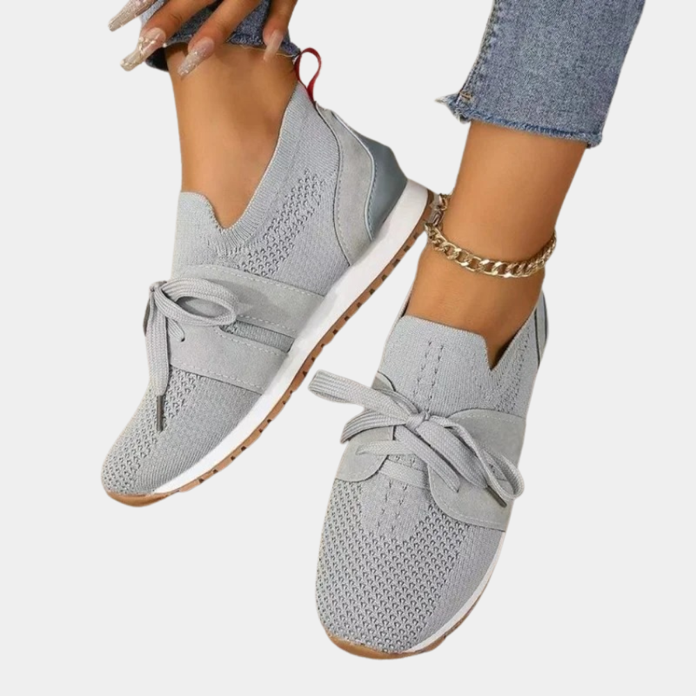 Chaussures de sport grises pour femme, design moderne avec lacets, semelle blanche antidérapante, portées avec un jean et un bracelet doré.
