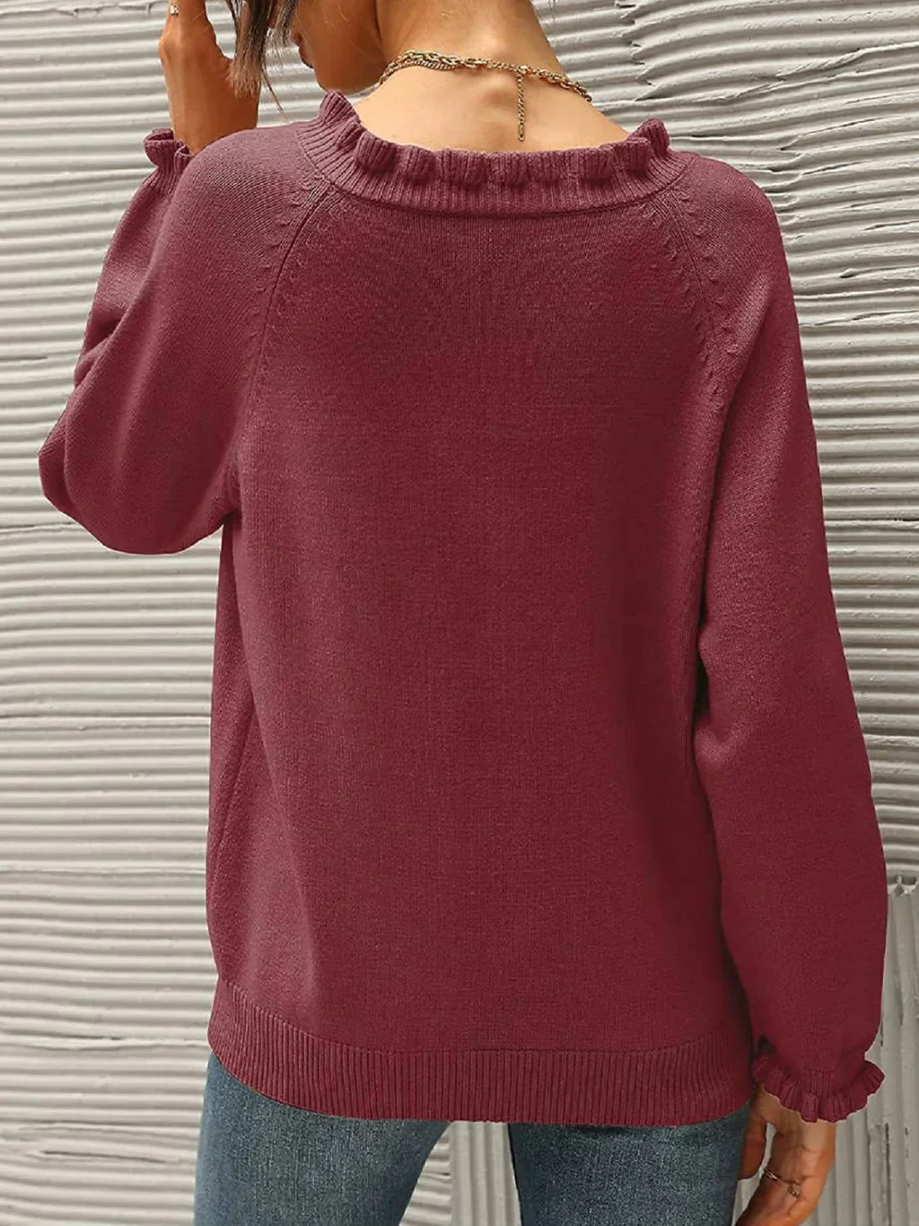 Femme portant un pull bordeaux à manches longues, col à volants, devant un mur texturé gris. Mode automne, style décontracté, tendance féminine.