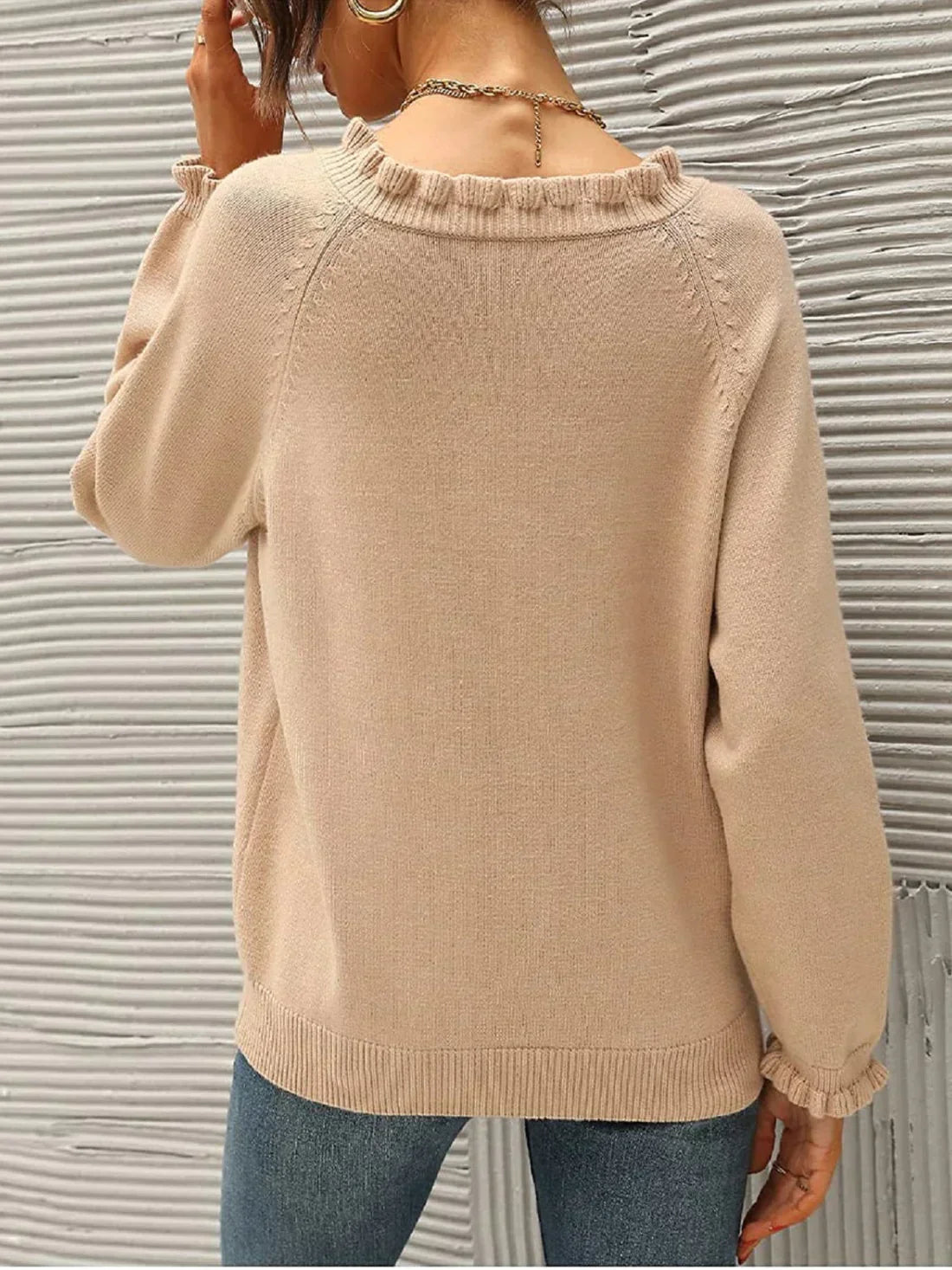 Femme portant un pull beige à manches longues avec col à volants, de dos, devant un mur texturé gris. Mode automne, style décontracté.