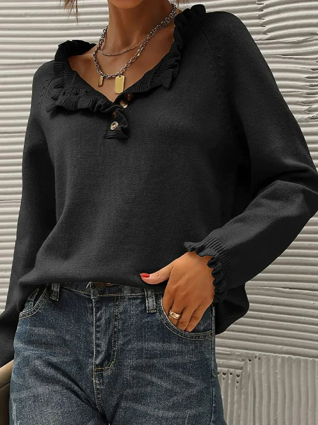 Femme portant un pull noir à volants avec col en V et boutons, associé à un jean bleu. Style chic et décontracté, mode automne-hiver tendance.