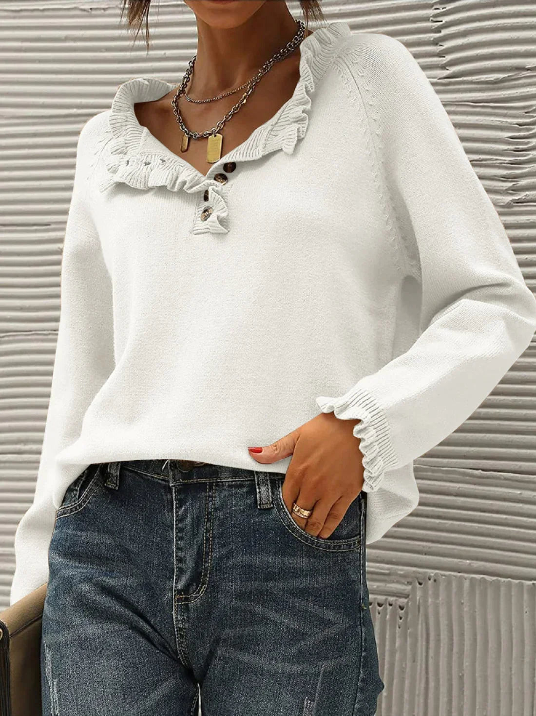 Femme portant un pull blanc à volants avec col en V, boutons décoratifs et manches longues, associé à un jean bleu. Mode automne-hiver élégante.
