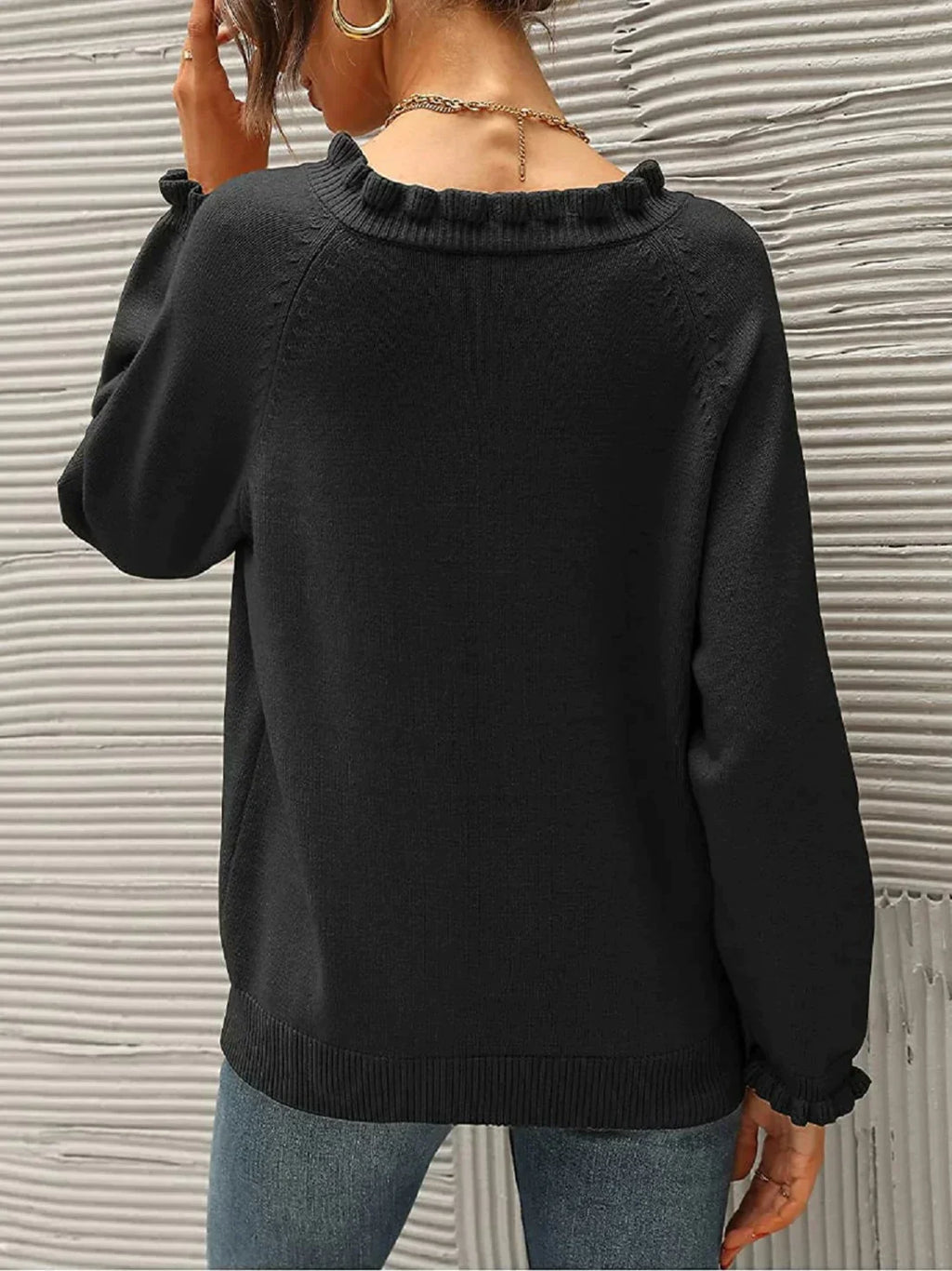 Femme portant un pull noir à col rond avec manches longues, dos tourné, devant un mur texturé gris. Mode automne, style décontracté, tendance.