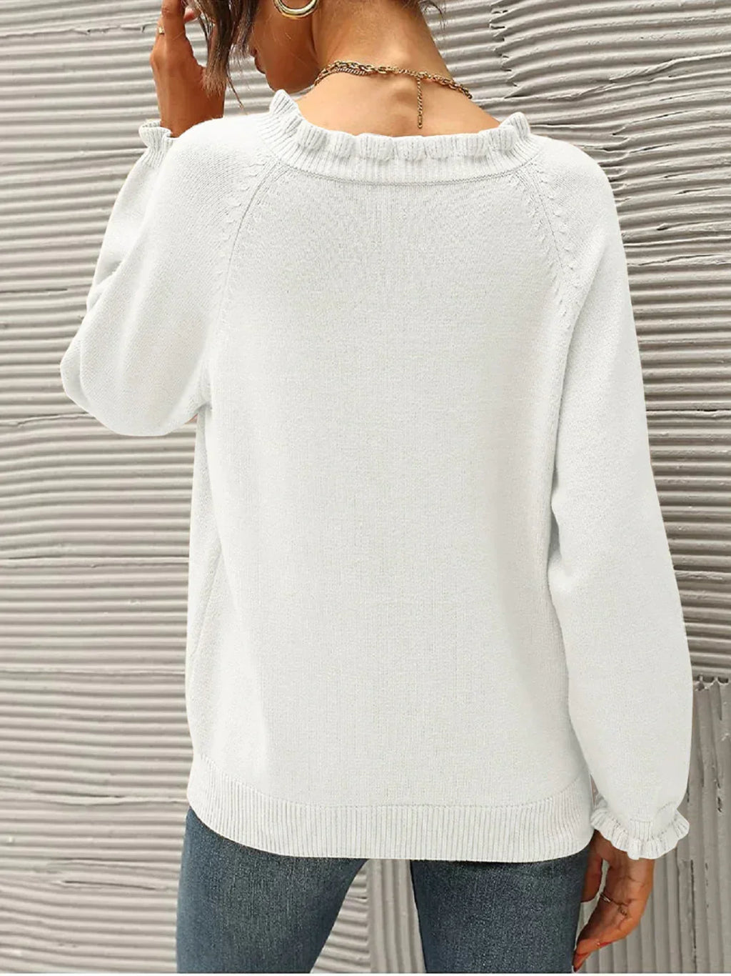 Femme portant un pull blanc en tricot, manches longues, devant un mur texturé gris. Mode automne, style décontracté, tendance féminine.
